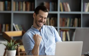 Excited man wearing glasses celebrating success, reading good news in email, happy overjoyed businessman looking at laptop screen, showing yes gesture and laughing, sitting at work desk